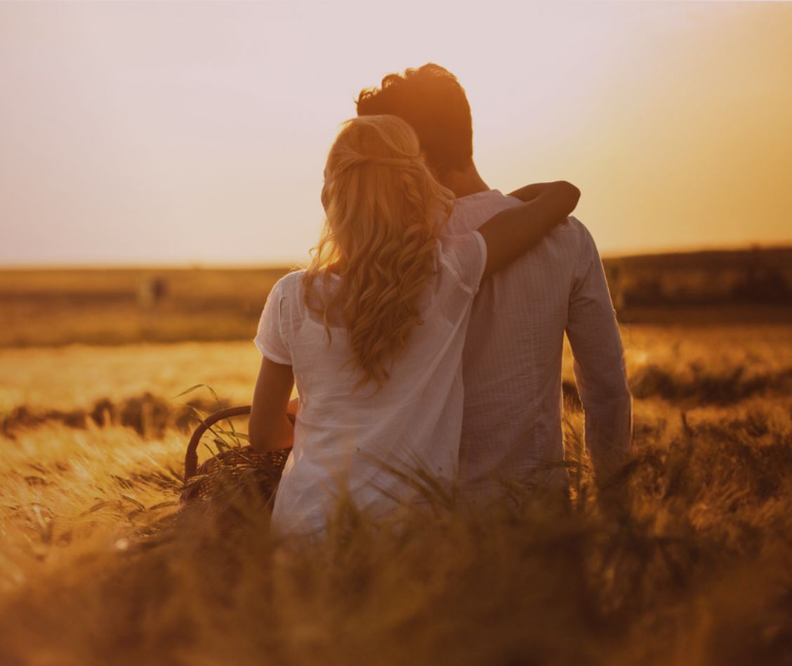 Young husband and wife sit together in the grass, the wife's arm around the husband's shoulder.